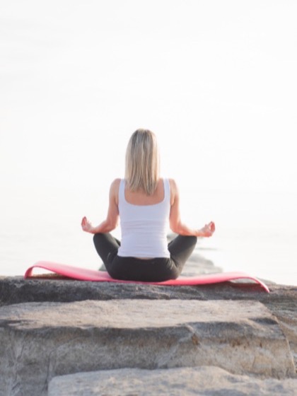 A person sitting cross-legged on a rock at the shoreline, facing a pale open sky.